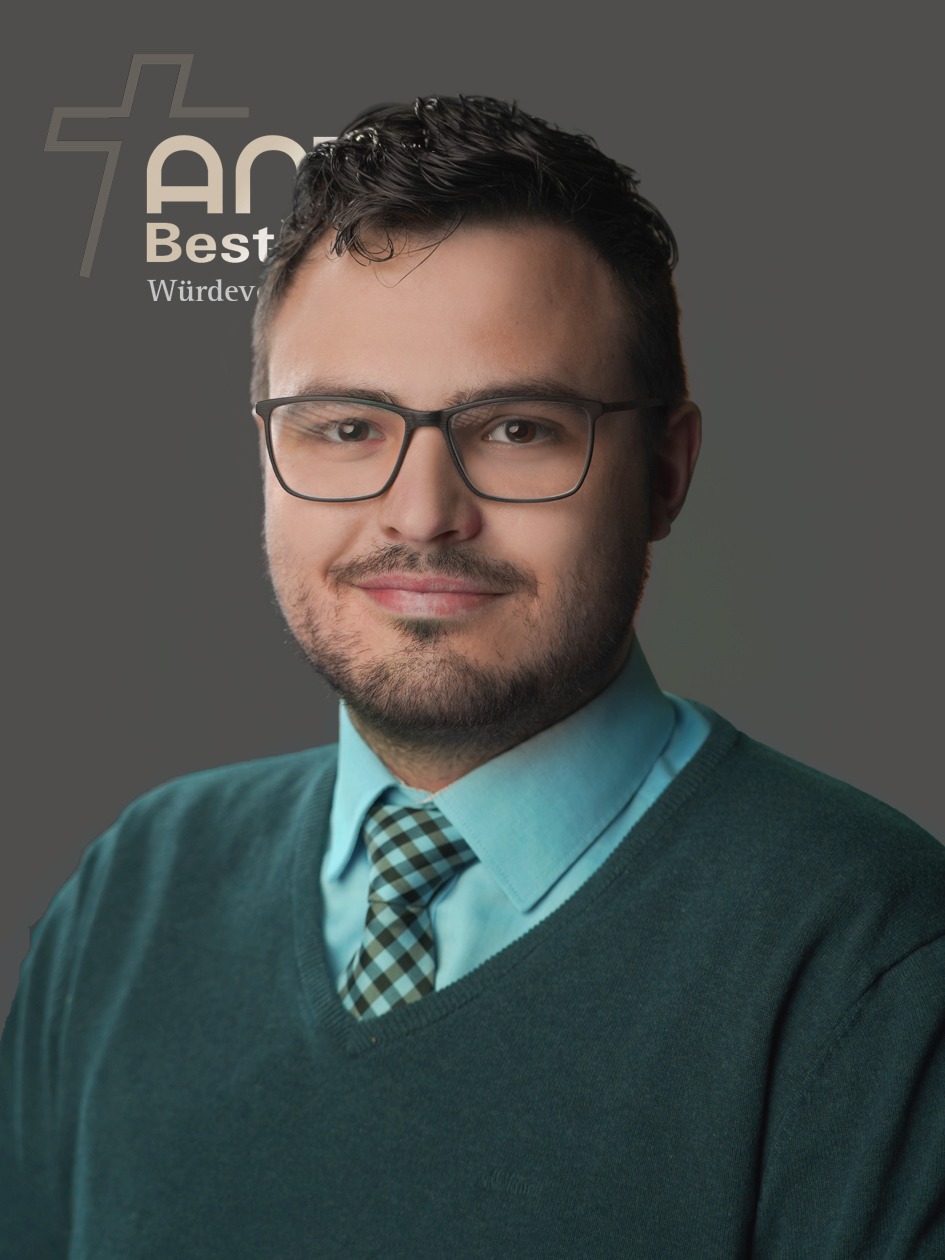 Ein Mann mit Brille, Bart und Schnurrbart steht vor einem grauen Hintergrund. Er trägt einen grünen Pullover und eine karierte Krawatte. Sein ernster Gesichtsausdruck und seine Kleidung lassen vermuten, dass er einer Bestattung beiwohnt, und verleihen der Szene einen Hauch nachdenklicher Selbstbesinnung. Bestattungsinstitut