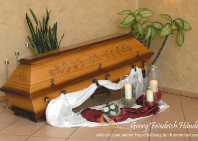 Ein wundersch&ouml;n gearbeiteter Holzsarg mit floralen Schnitzereien, umgeben von Kerzen, Stoff und Pflanzen, ziert den beigefarbenen Raum &ndash; eine heitere Szene, die eine zeitlose Bestattungstradition widerspiegelt.