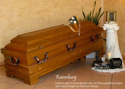 Ein kunstvoller Holzsarg mit bronzenen Akzenten, verziert mit floralen Schnitzereien und einem Blumenstrau&szlig;. In der N&auml;he flackert eine Kerze, w&auml;hrend eine kleine Metallbahn und die w&uuml;rdevolle Atmosph&auml;re diese feierliche Bestattung widerspiegeln.