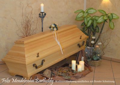 Ein Holzsarg mit kunstvollen floralen Schnitzereien steht inmitten leuchtender Kerzen und &uuml;ppiger Pflanzen in einem ruhigen Raum und erinnert an die zeitlose Eleganz Felix Mendelssohn Bartholdys. Diese ergreifende Szene spiegelt eine w&uuml;rdevolle Bestattung wider, die Leben und Verm&auml;chtnis ehrt.