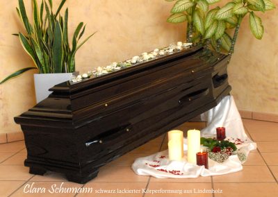 Ein eleganter schwarzer Sarg mit Blumenarrangement, umgeben von Kerzen und Pflanzen auf dem gefliesten Boden, schafft eine elegante Bestattungsatmosph&auml;re.