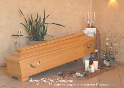 Ein mit Kerzen, Pflanzen und Blumen geschm&uuml;ckter Holzsarg ruht anmutig auf einem gefliesten Boden. Eine Atmosph&auml;re der Ruhe umgibt die Szene, mit der dezenten Inschrift &bdquo;Georg Philipp Telemann&ldquo; &ndash; eine heitere Hommage an eine w&uuml;rdevolle Bestattung.