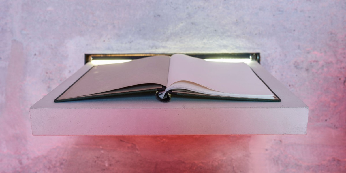 Ein offenes Buch liegt auf einem minimalistischen Regal, sanft hinterleuchtet vor einer strukturierten Wand. Diese ruhige Umgebung kontrastiert mit dem Gedanken an Bestattung und l&auml;dt in ihrer friedlichen Atmosph&auml;re zum Nachdenken und Reflektieren ein.
