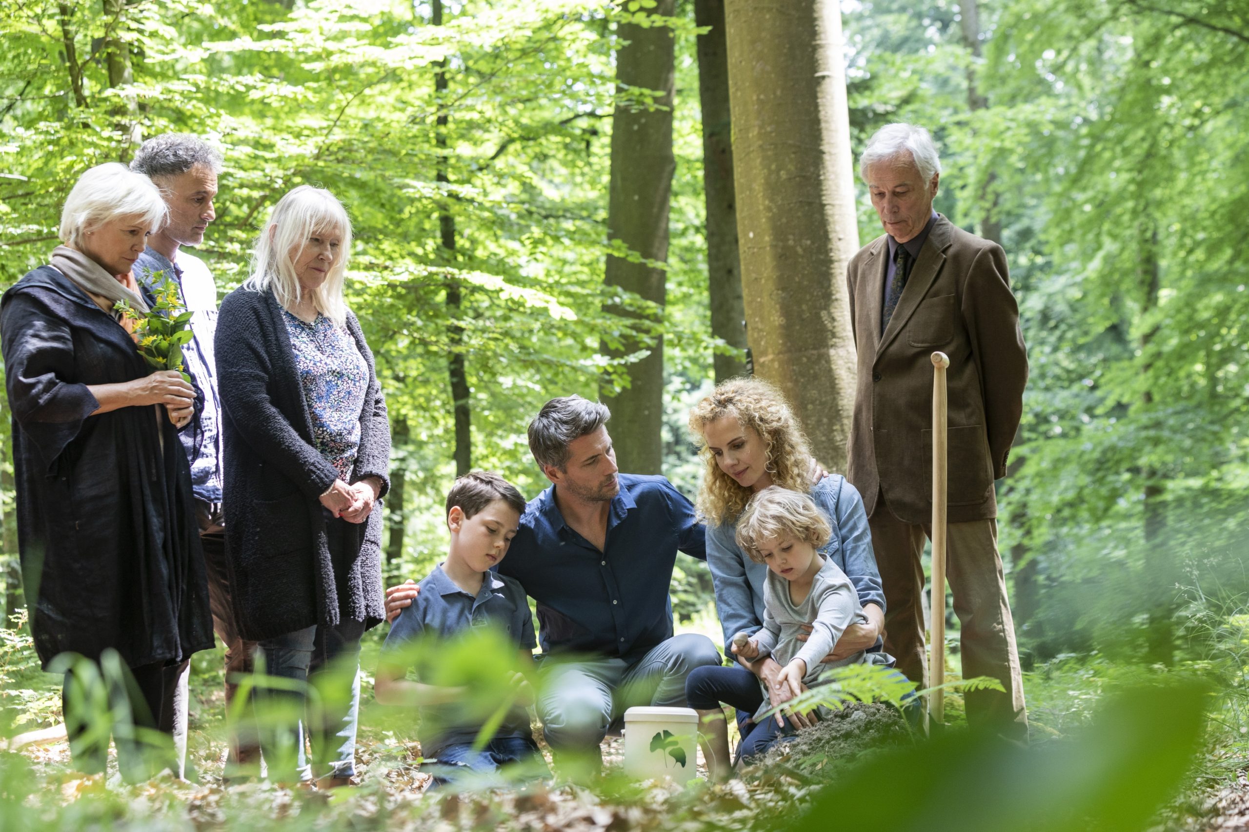 Eine Gruppe von Menschen versammelt sich im Wald feierlich um einen Baumstumpf und nimmt an einem d&uuml;steren Ritual teil, das an eine Bestattung erinnert.