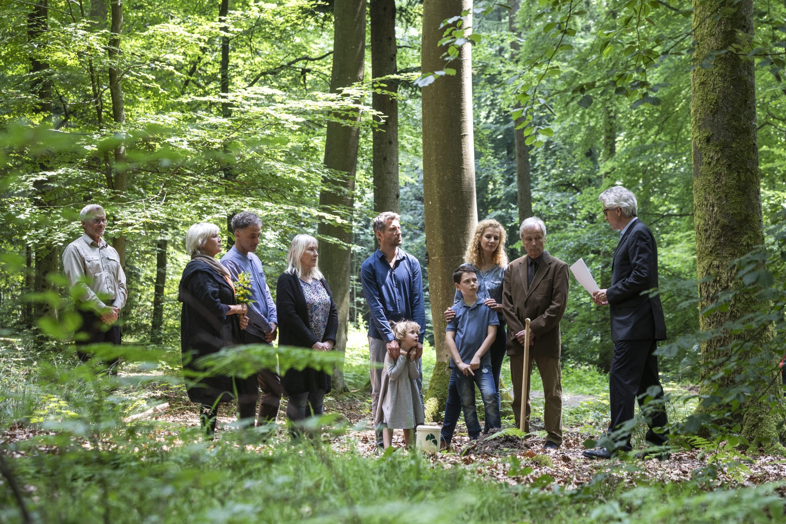 In einer ruhigen Waldlandschaft versammelten sich die Menschen zu einer herzlichen Bestattungszeremonie, w&auml;hrend ein Mann respektvoll zu der aufmerksamen Gruppe sprach.