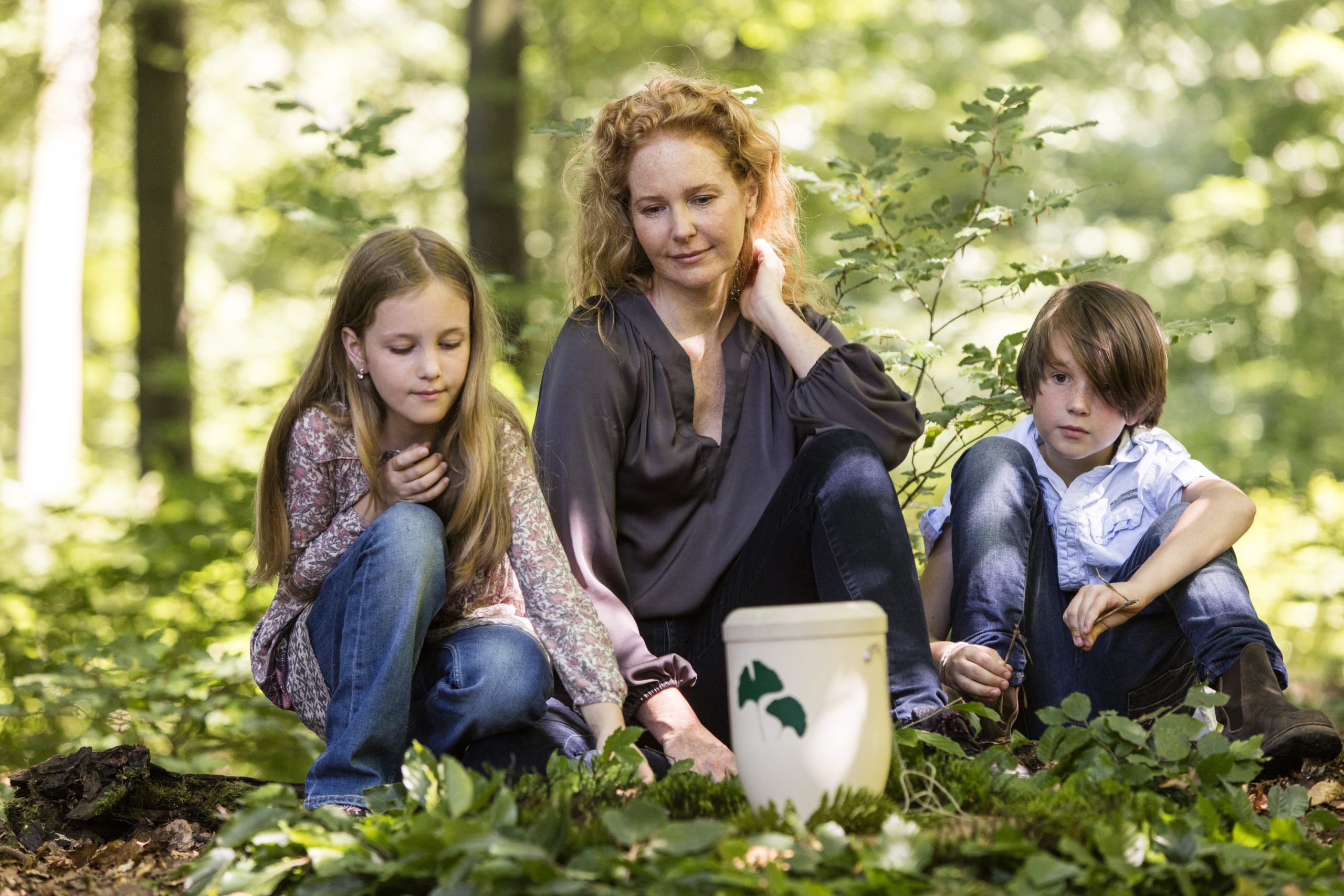 Eine Frau und zwei Kinder sitzen ruhig auf dem Waldboden neben einem wei&szlig;en Eimer mit gr&uuml;nem Blattmuster. Die friedliche Szene erinnert an die stille Feierlichkeit, die oft mit Bestattungszeremonien verbunden wird, da sie von der Ruhe der Natur umh&uuml;llt wird.