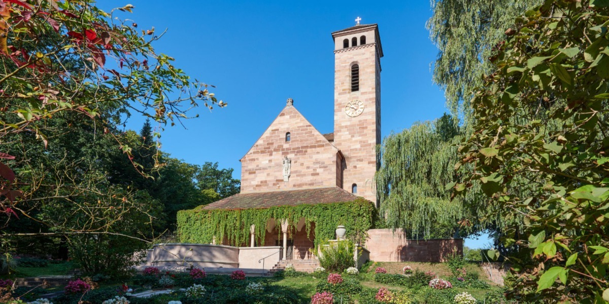 Eine historische Steinkirche mit einem Glockenturm steht anmutig, umgeben von &uuml;ppigem Gr&uuml;n und leuchtenden Blumen unter einem klaren blauen Himmel und bietet eine heitere Kulisse f&uuml;r Bestattungen.