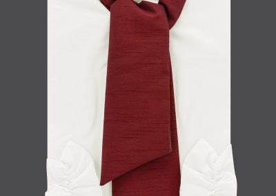 Auf grauem Hintergrund ist ein gefaltetes wei&szlig;es Hemd mit roter Krawatte und Kragen sowie wei&szlig;en R&uuml;schenmanschetten zu sehen &ndash; ein klassisches Ensemble, das an die formelle Kleidung einer Bestattung erinnert.